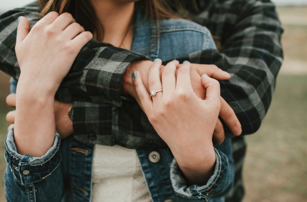 Woman wearing engagement ring being embraced from behind by her male partner.