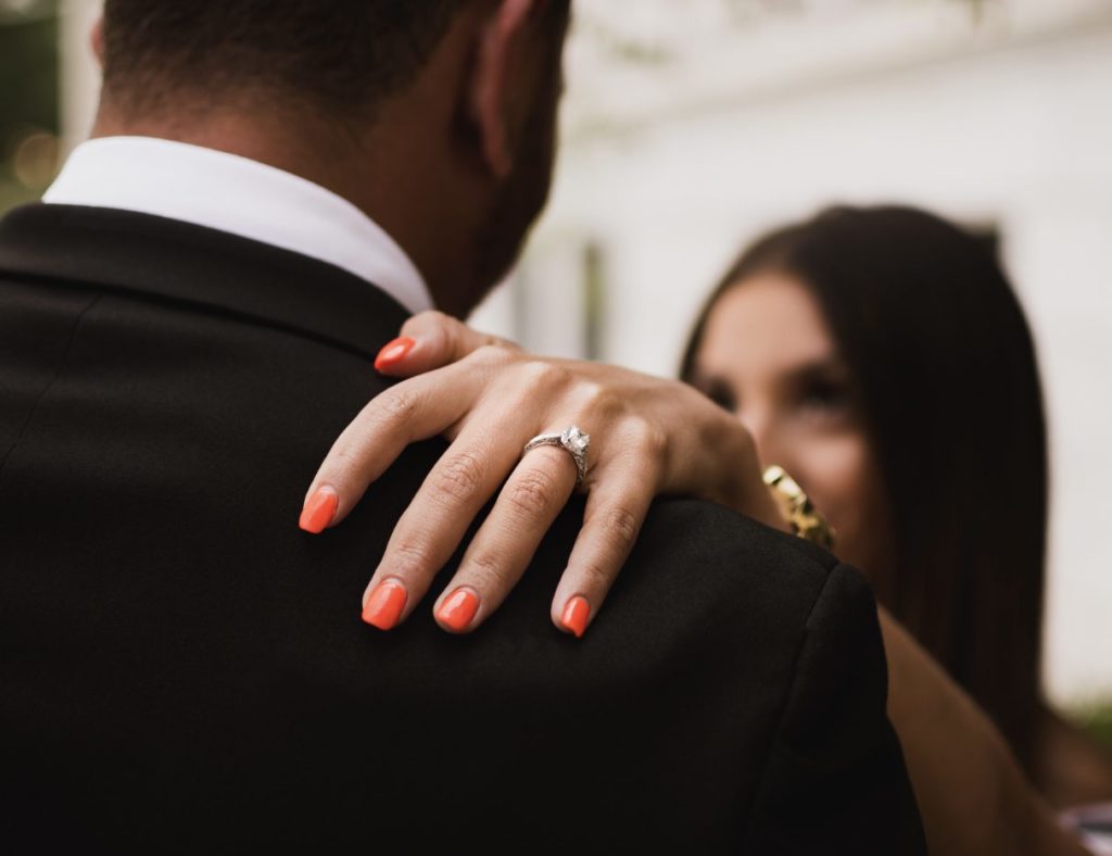 Closeup of woman's hand resting on her partner's shoulder and wearing a diamond engagement ring.